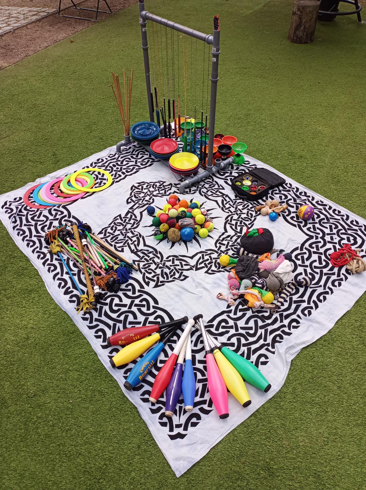Mitmachzirkus25 Konzertplatz Auf einer großen Decke liegen verschiedene Zirkusrequisiten: Bälle, Keulen, Ringe, Diabolos, Flowersticks, Jojos, Poi und Teller.